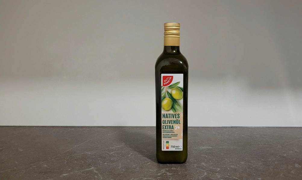 Eine Flasche getestetes Olivenöl von vorne auf einer dunklen Fläche vor einer hellen Wand.