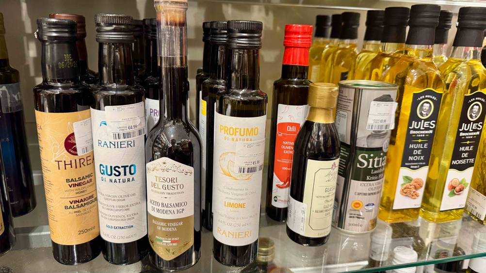 Mehrere Flaschen des Olivenöl mit Zitrone im Test in einem Verkaufsregal neben anderen Produkten.