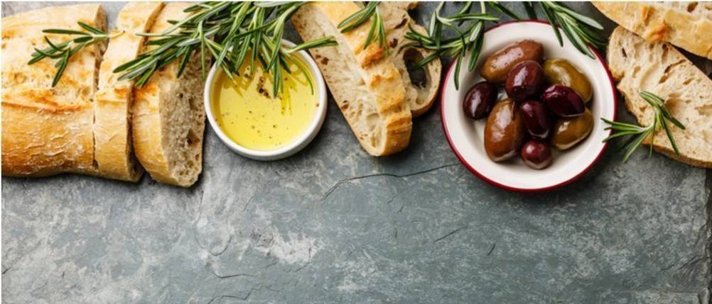 Bertolli-Olivenöl-Test: Auf einer Küchenfläche liegt ein aufgeschnittenes Baguette. Daneben stehen zwei Schälchen mit Oliven und Olivenöl. Das Baguette ist mit Rosmarin garniert. 