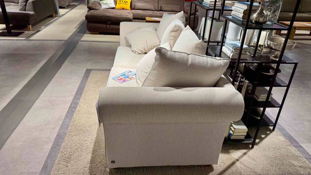 Im Test: Ein 3-Sitzer Sofa mit hellem Bezug steht vor einem schwarzen Regal im Ausstellungsbereich eines Möbelhauses.