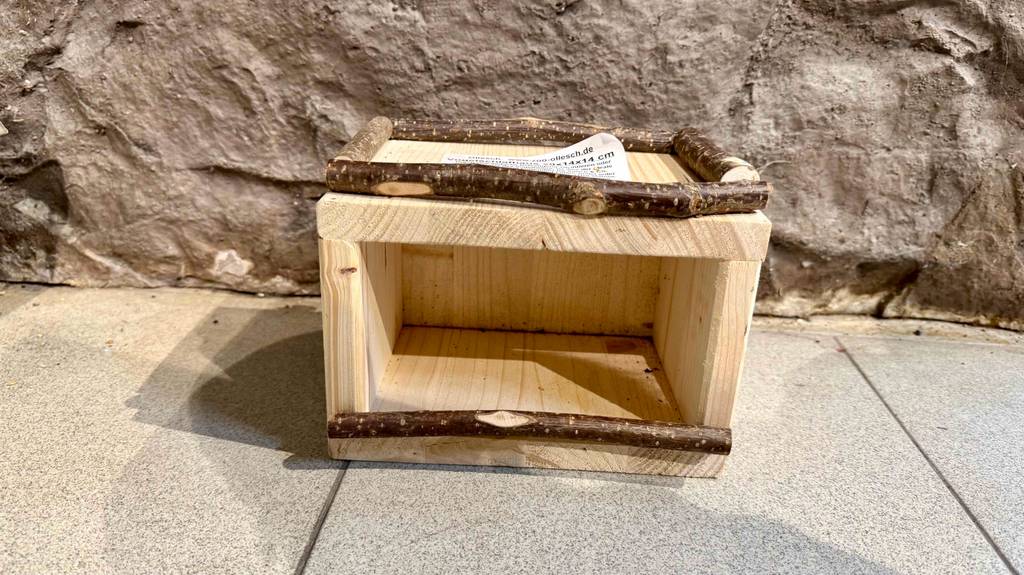 Ein Vogelhaus aus Holz steht zum testen vor einer Steinwand.