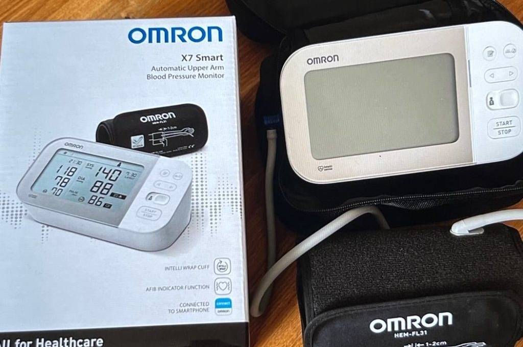 Oberarm-Blutdruckmessgerät-Test: Omron-Modell neben seiner Originalverpackung mit Produktbeschreibung auf einem Holzuntergrund.
