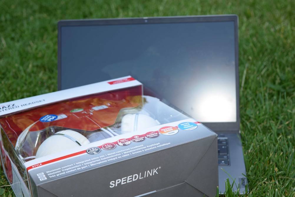 Im Test: Ein originalverpackter On-Ear-Kopfhörer von schräg unten vor einem Laptop auf einer Wiese.