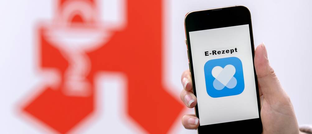 Online-Apotheken getestet: Eine Person hält vor einem Apotheken-Logo ein Smartphone mit einem E-Rezept darauf.