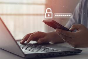 Online-Banking-Software-Test: Eine Person sitzt an einem Laptop und hält ein Smartphone in der Hand, über dem eine Grafik schwebt, die eine Passwort-Zeile darstellt.
