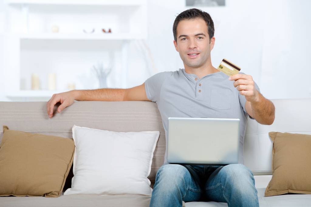 Die beste Banking-Software im Vergleich: Ein Mann sitzt mit einem Laptop auf den Beinen auf der Couch und präsentiert seine Bankkarte.