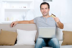 Online-Banking-Software-Test: Ein Mann sitzt mit einem Laptop auf den Beinen auf der Couch und präsentiert seine Bankkarte.