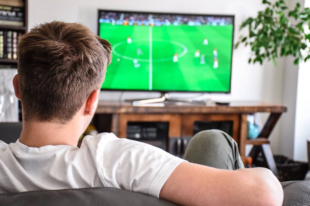 Ein Mann sitzt auf der Couch und schaut über seinen Fernseher eine Fußball-Übertragung.