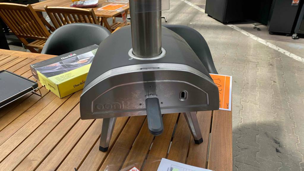 Detailansicht von hinten auf einen anthrazitfarbenen Ooni Garten-Pizzaofen im Test, der mit Holzpellets beheizt wird und auf einem im Baumarkt stehenden Holz-Gartentisch ausgestellt ist.