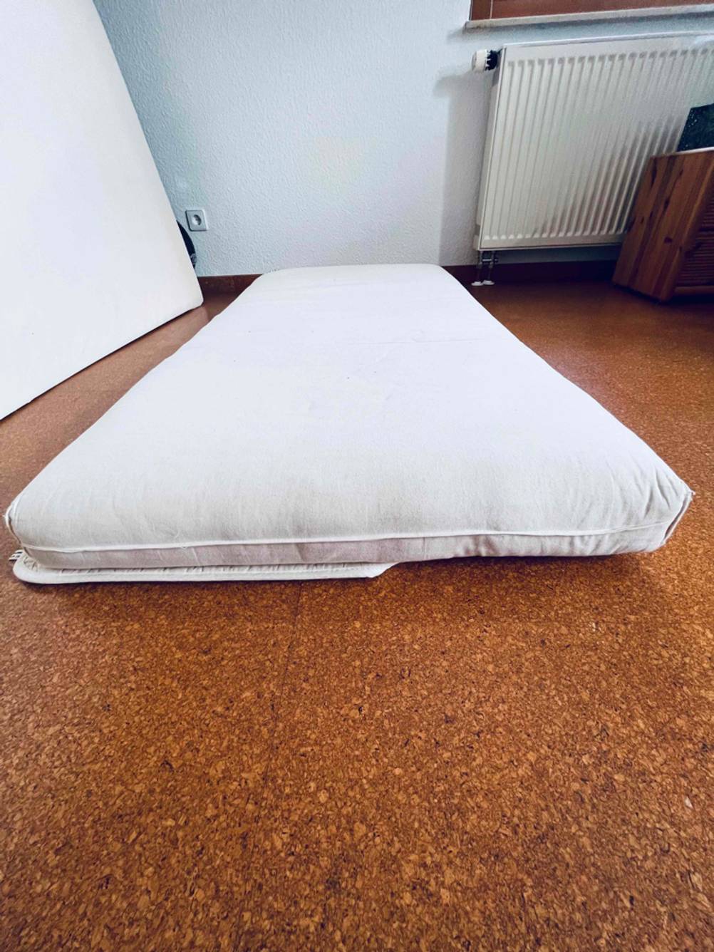 Weiße Kindermatratze im Test liegt auf einer Matratzenunterlage auf einem braunen Fußboden im Wohnbereich.