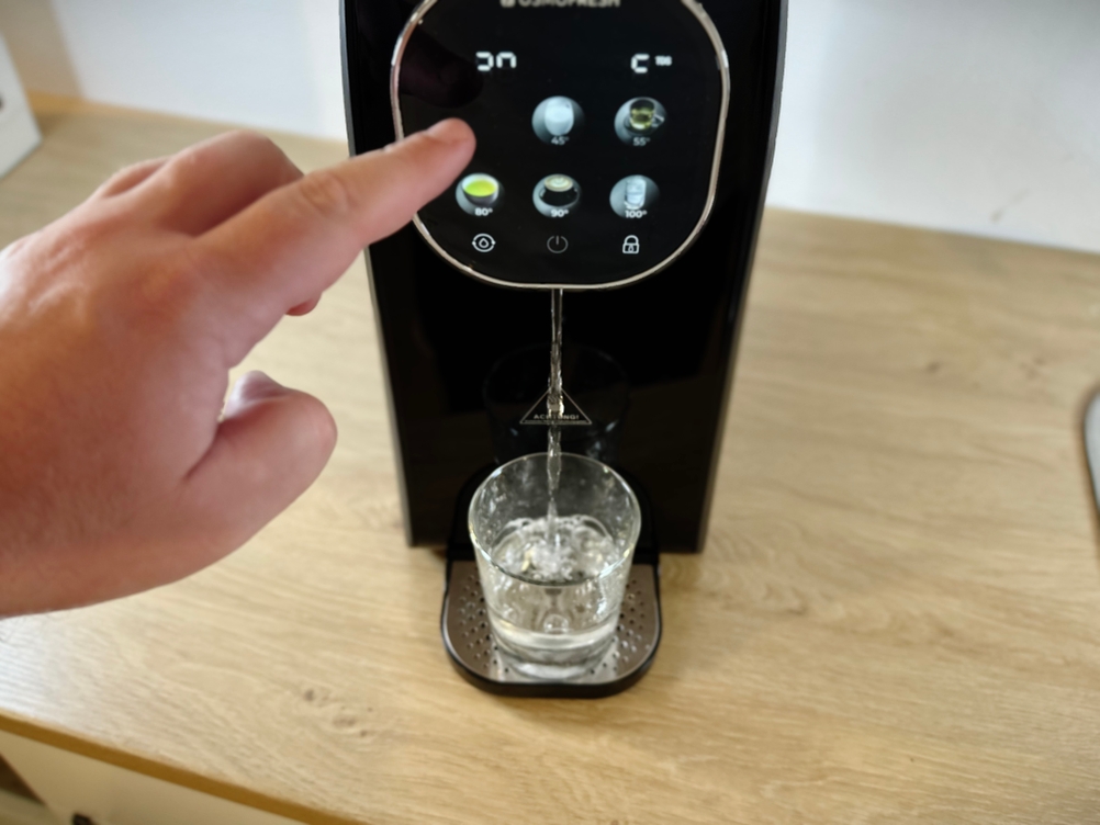 Ein Finger tippt auf das Display der Osmofresh Quella Pro Black, während ein Glas mit Wasser befüllt wird. Das Gerät gibt gerade Wasser aus.