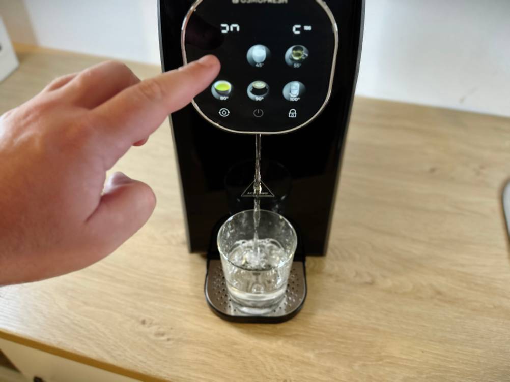 Ein Finger tippt auf das Display der Osmofresh Quella Pro Black, während ein Glas mit Wasser befüllt wird. Das Gerät gibt gerade Wasser aus.