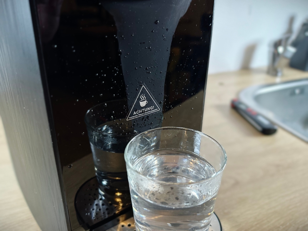 Wassertropfen an der schwarzen Front der Osmofresh Quella Pro Black, im Vordergrund steht ein Glas mit gefiltertem Wasser. Das Symbol „Achtung heiß“ ist sichtbar.