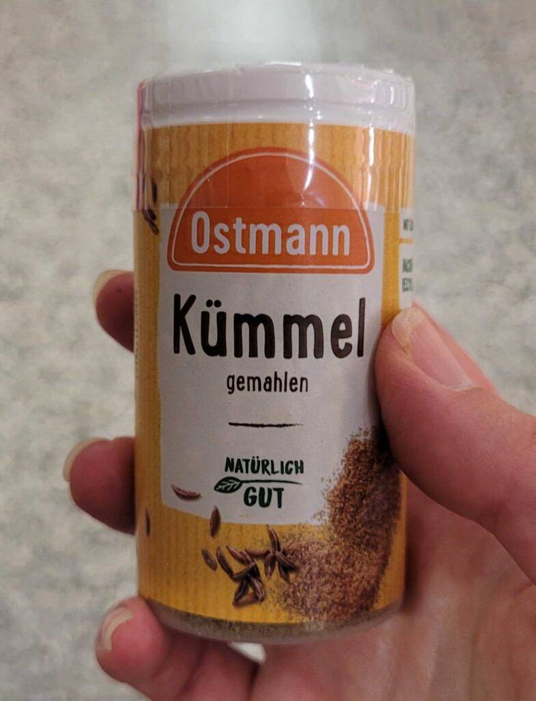 Eine Hand hält eine Dose mit gemahlenem getesteten Kümmel vor einem grauen Hintergrund.