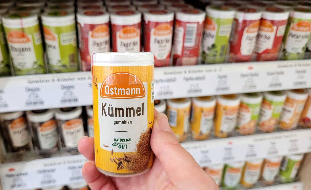 Kümmel-Test: Eine Hand hält eine Dose mit gemahlenem Kümmel vor einem Gewürzregal.