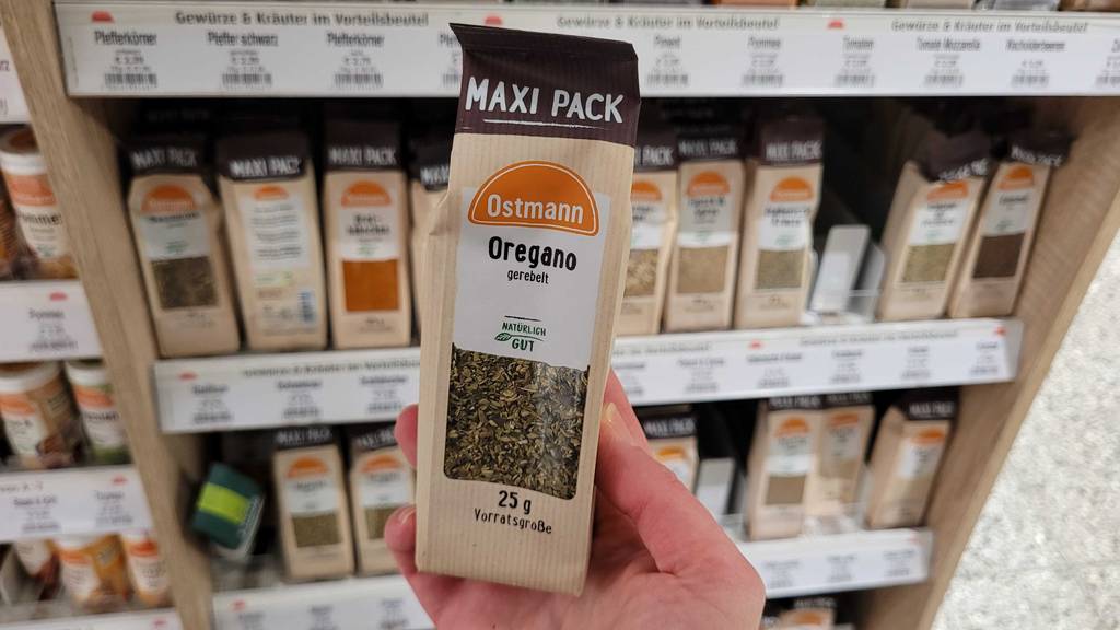 Eine Hand hält ein 25-g-Maxi-Pack mit getestetem Oregano vor einem Gewürze-Regal im Supermarkt.
