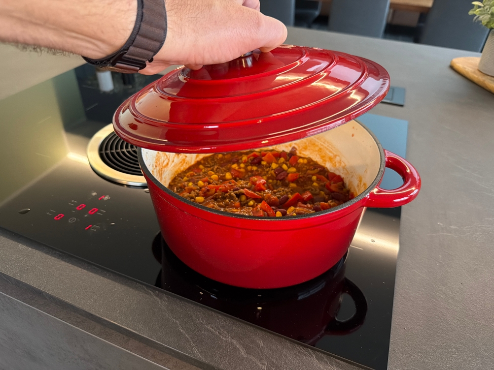Eine Hand hebt den Deckel von einem roten Schmortopf auf dem Herd ab.