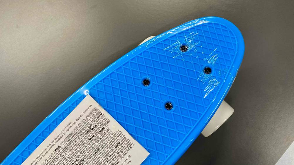 Detailaufnahme der in Zellophan eingeschweißten Spitze eines blauen Skateboards für Kinder im Test auf grauem Untergrund.