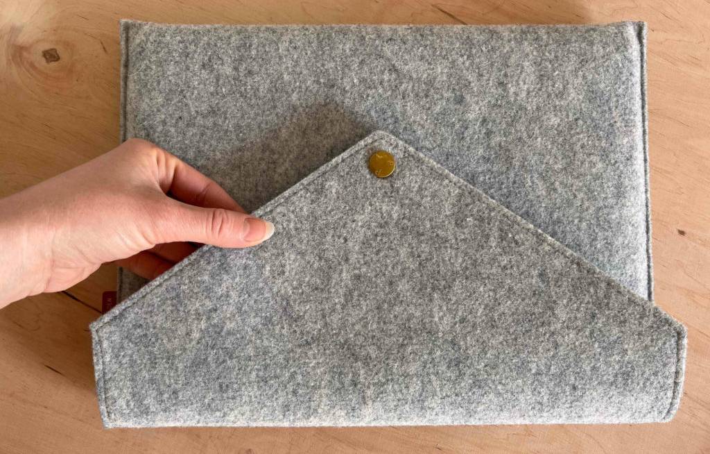 Aufnahme von oben auf eine Hand, die den Brief-Verschluss inklusive Druckknopf einer grau melierten Woll-MacBook-Tasche auf Holz berührt.