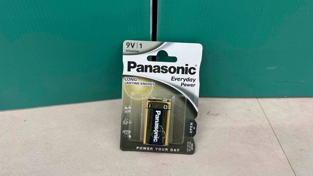Eine getestete 9V-Batterie steht in der Verpackung vor einer grünen Wand.
