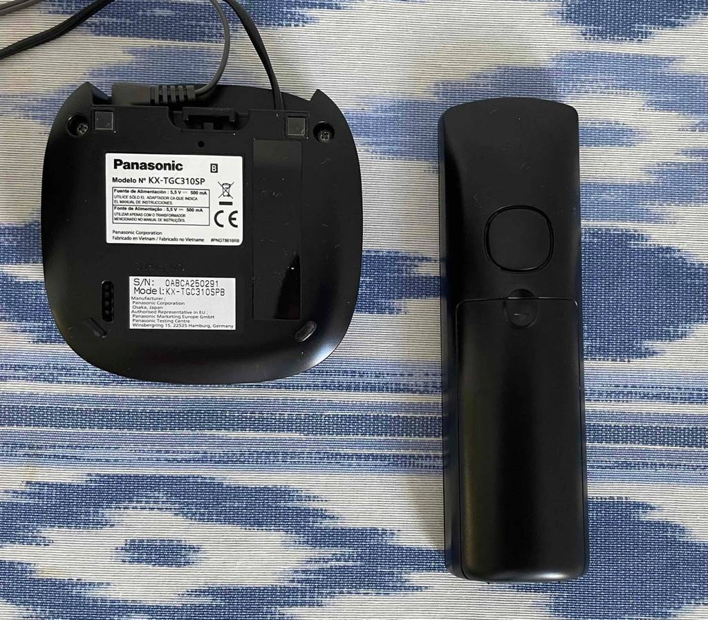Ein getestetes schnurloses Telefon der Marke Panasonic liegt von unten betrachtet, inklusive Produktinformation auf der Basis-Station, auf einem blau-weißen Gewebe.