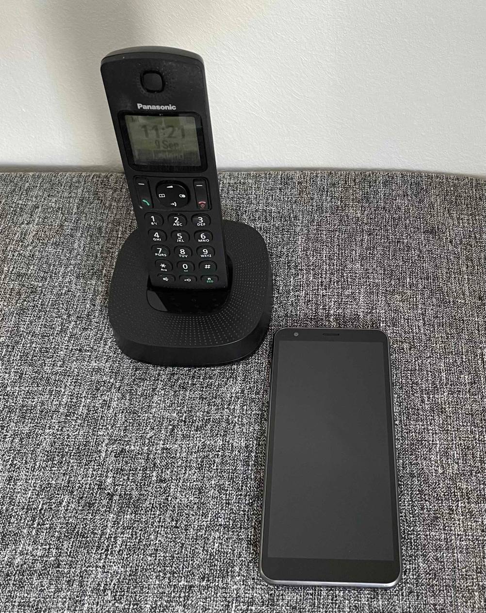 Im Test: Schnurloses Telefon in Schwarz der Marke Panasonic befindet sich auf seiner Basis neben einem Smartphone auf grau-meliertem Stoff.