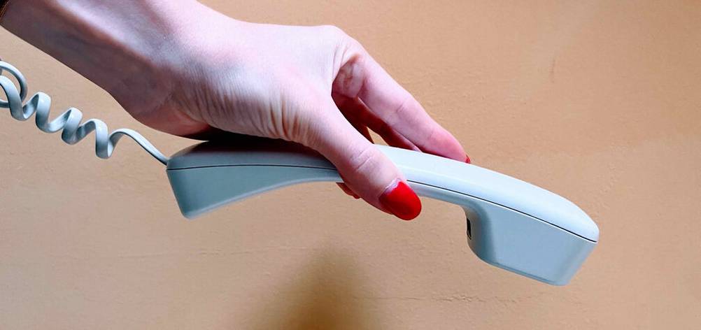 Schnurlostelefon-Test: Seitenansicht eines weißen Telefonhörers inklusive Kabel in einer Hand vor beigefarbener Wand.