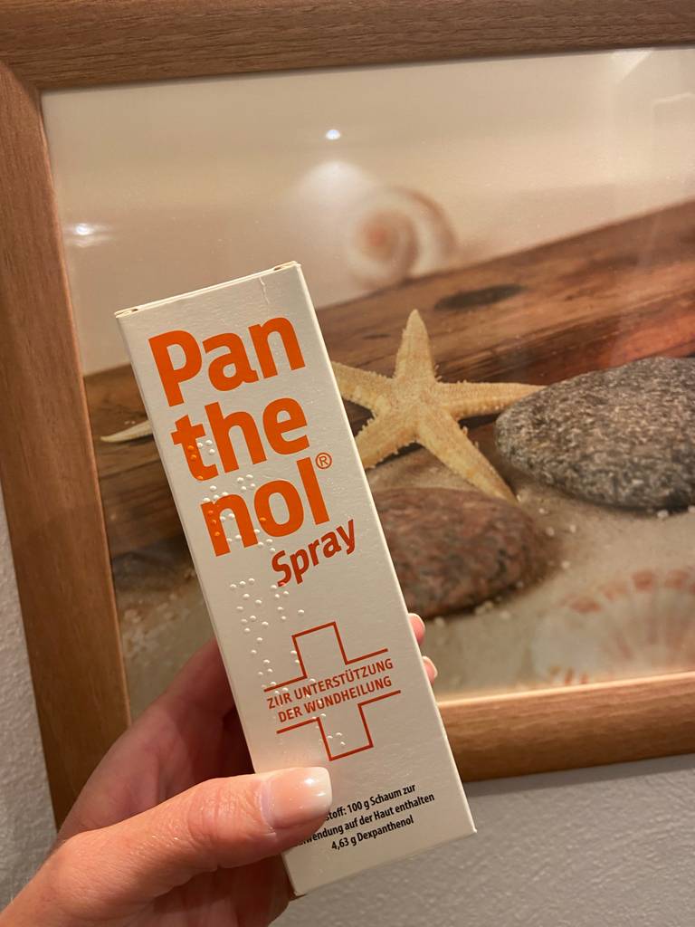Eine Hand hält eine Packung Panthenol-Spray zum testen vor einem Dekobild.