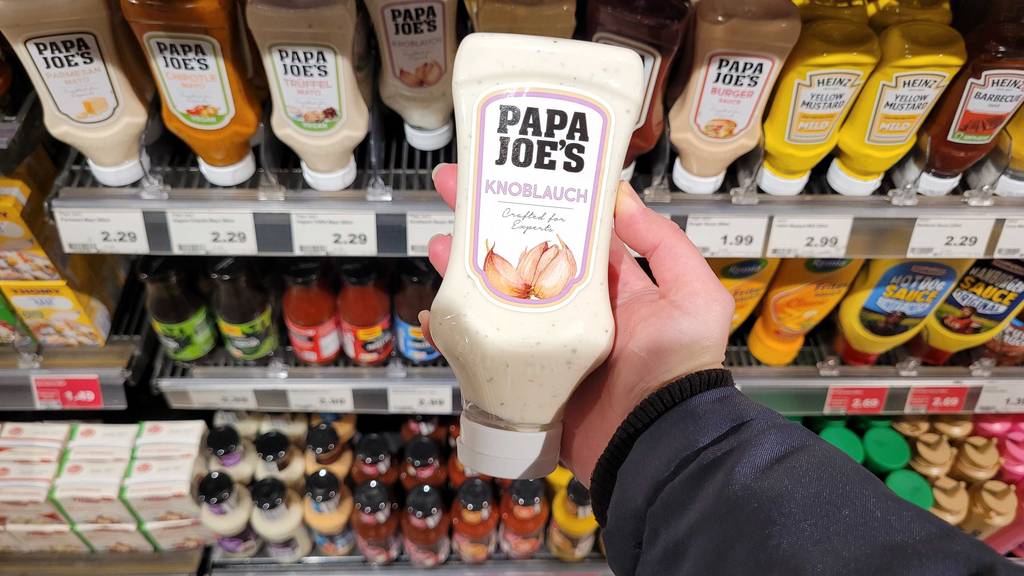 Eine Hand hält eine Quetschflasche Knoblauch-Sosse zum testen vor einem Verkaufsregal.