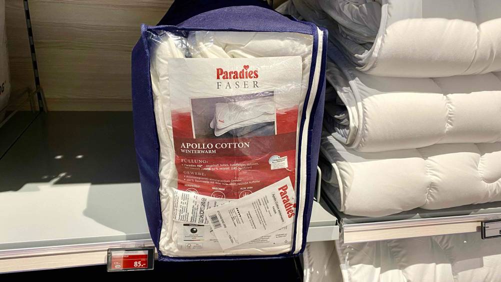 Eine blaue Verpackung inklusive einer Paradies-Thermobettdecke im Test steht inklusive Produktbeschreibung und Zertifikaten auf einem Verkaufsregal neben ausgepackten weißen Bettdecken.