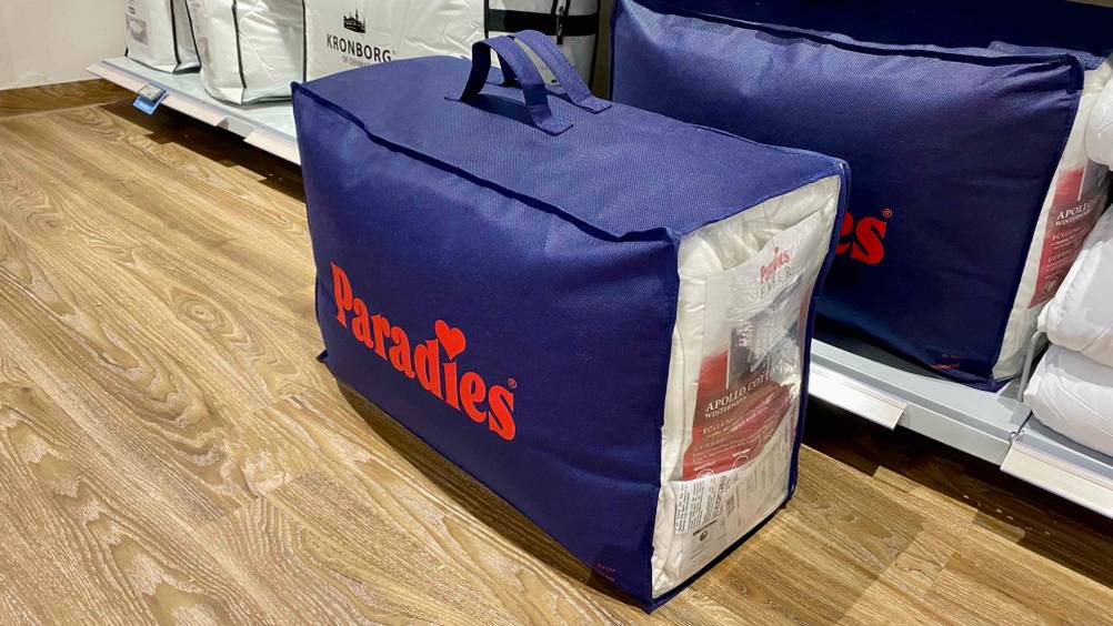Ansicht einer blauen Verpackung mit Griffen, die eine Paradies Thermobettdecke im Test enthält, steht auf einem Holzboden vor einem Verkaufsregal mit weiteren verpackten Bettdecken.