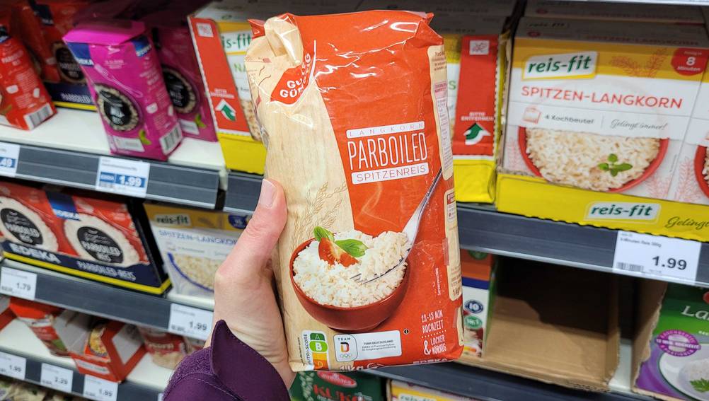 Eine Hand hält eine Packung des getesteten Parboiled-Reis vor ein Regal.