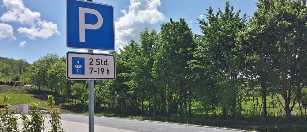 Parkscheiben-Test: Parkplatzschild mit Höchstparkdauerzusatz auf einem leeren Parkplatz, umgeben von Bäumen 
