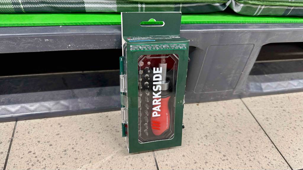 Ratschenschraubendreher im Test: Ein Modell mit orangem Griff und mehreren Aufsätzen steht innerhalb seiner Aufbewahrungsbox vor einem Verkaufsregal.