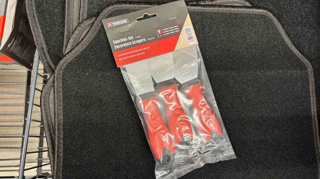 Spachtel im Test: Drei verpackte Spachtel mit orangem Griffen liegen auf einer schwarzen Matte.