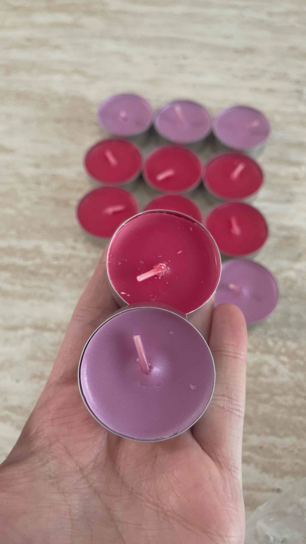 Eine Hand hält zwei getestete Partylite-Kerzen über mehreren malvenfarbigen und roten Teelichtern, die auf einer braunen Oberfläche stehen.