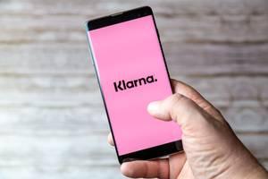 Pauschalreisen im Test: Ein Mann drückt mit dem Daumen auf ein Smartphone-Display mit dem Klarna-Logo.