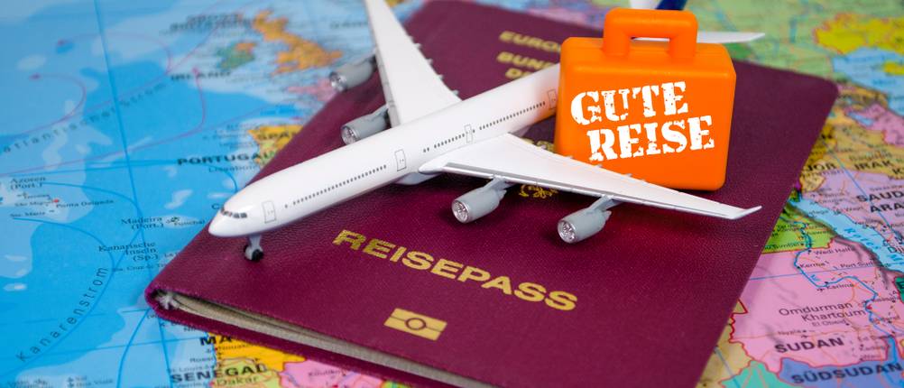 Pauschalreisen-Test: Auf einer Weltkarte liegen ein Reisepass, ein Spielzeugflugzeug und ein kleiner Koffer mit der Aufschrift 