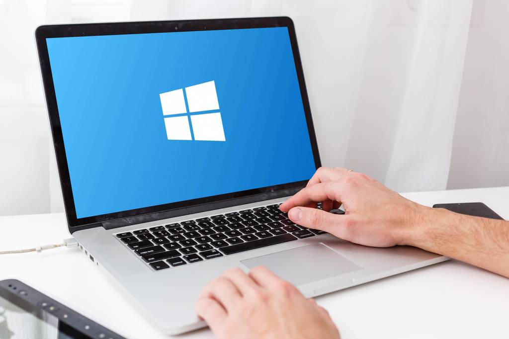 Die Hände eines Mannes liegen auf der Tastatur eines silbernen Laptops mit Windows-Logo im Bildschirm.