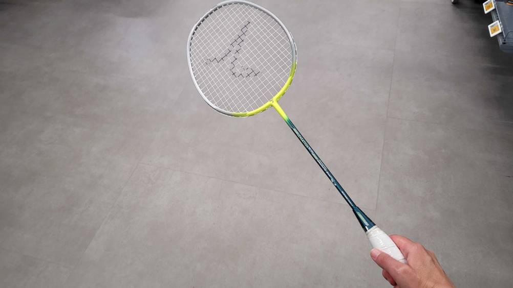 Eine Hand hält einen Badmintonschläger im Test vor einen grauen Boden.