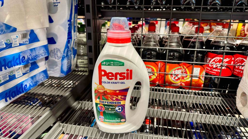 Eine Flasche getestetes Persil-Flüssigwaschmittel steht neben WC-Papier und vor Getränken in einem Gitterregal.