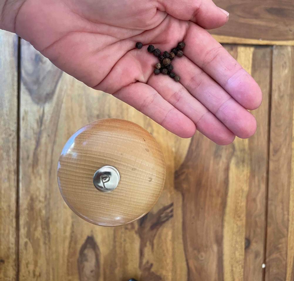 Eine getestete Holz-Pfeffermühle von oben, daneben eine Handfläche in der sich Pfeffer befindet.