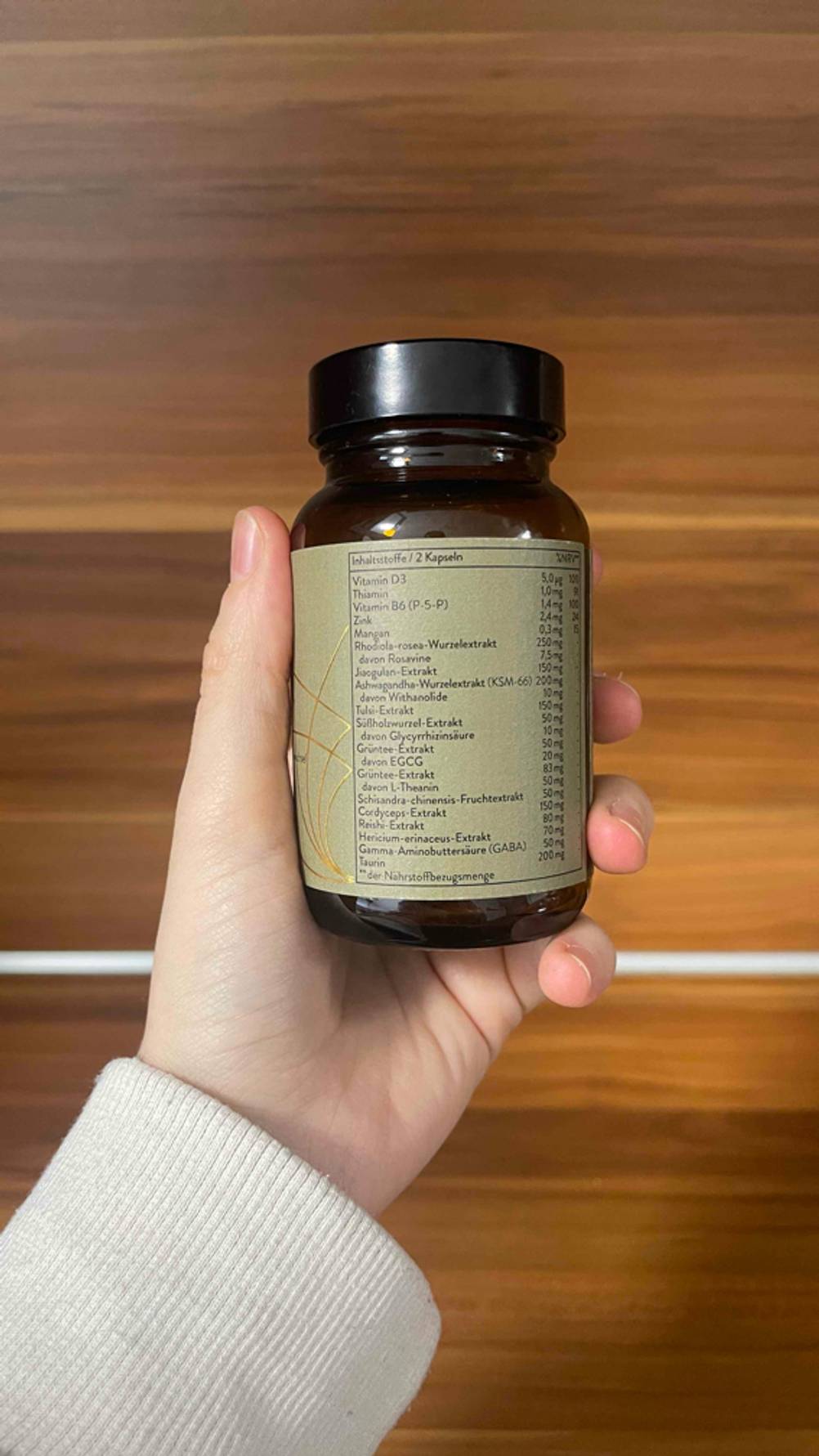 Eine Hand hält eine blickdichte Dose mit olivefarbenem Etikett inklusive der Produktinformationen vor einem Holzhintergrund. Darin befindet sich das pflanzliche Beruhigungsmittel im Test.