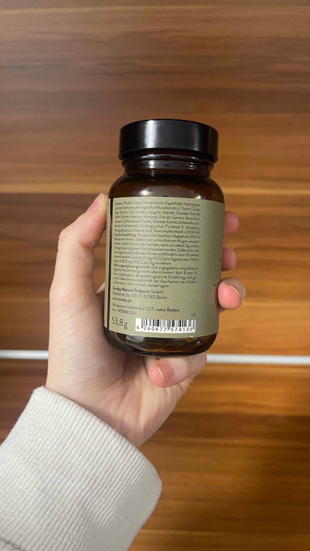 Eine Hand hält die blickdichte Dose mit olivefarbenem Etikett mit daruaf gedruckten Produktinformationen vor einer Holzfläche. In dem Braunglas befindet sich das getestete pflanzliche Beruhigungsmittel.