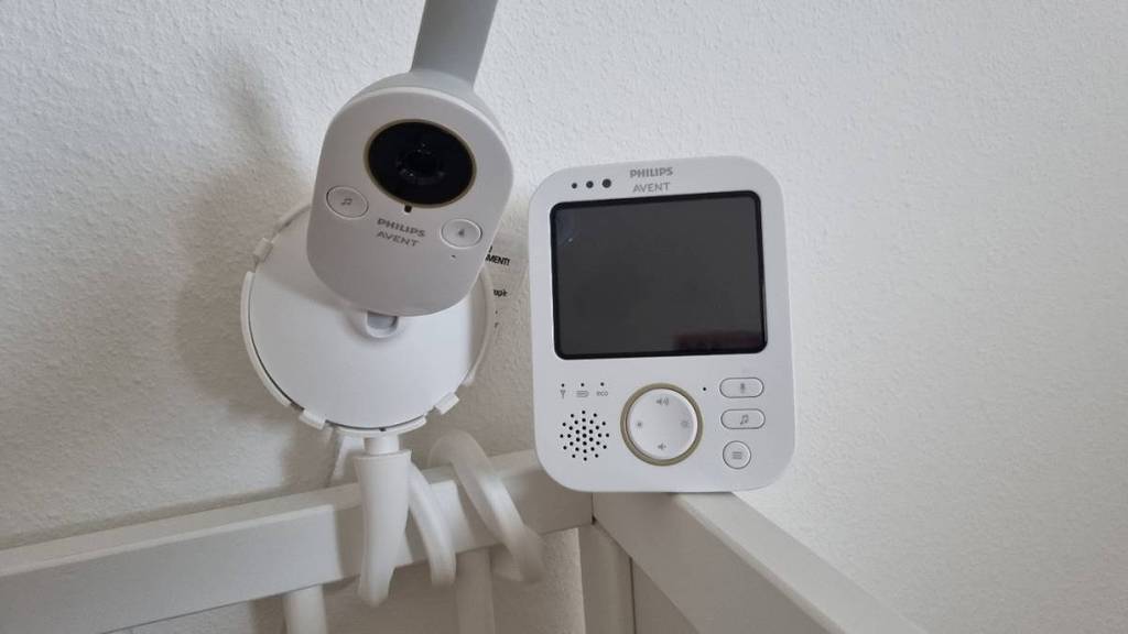 Philips-Avent-Baby-Kamera im Test: eine Kamera im weißen Design inklusive biegsamem Standfuß ist an einem weißen Kinderbett-Geländer befestigt. Daneben befindet sich das Bildschirm-Element.
