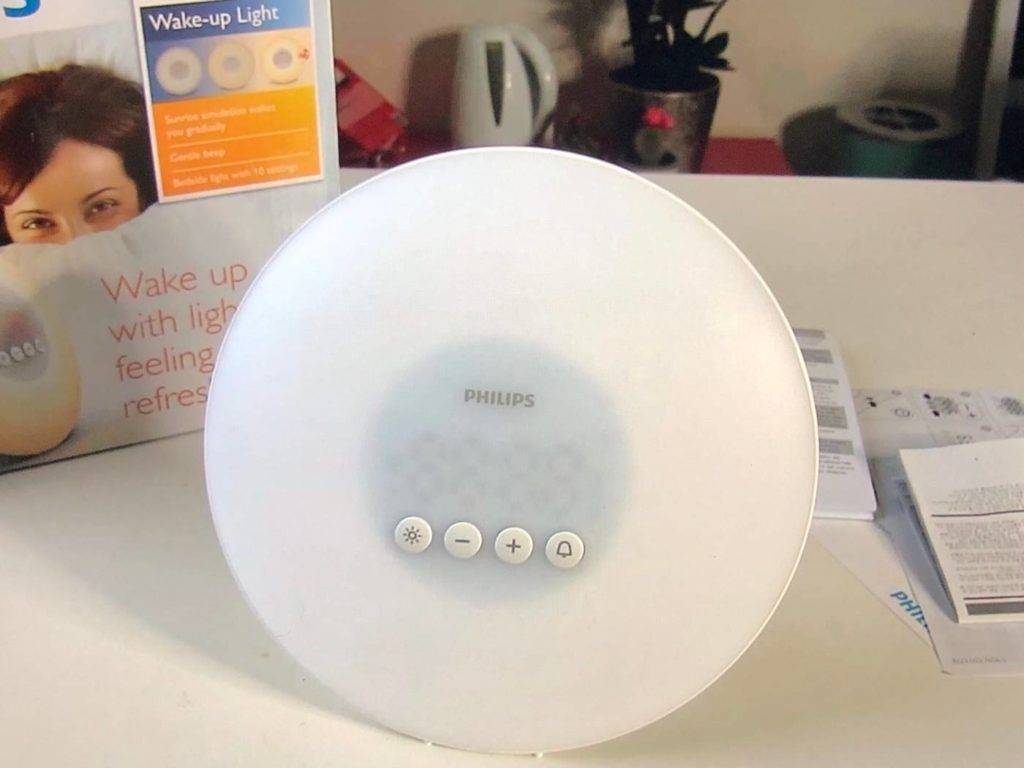 Lichtwecker im Test: Nahaufnahme eines runden, weißen Philips Lichtweckers. Er steht ausgeschaltet seitlich vor seiner Originalverpackung in einem häuslichen Ambiente.