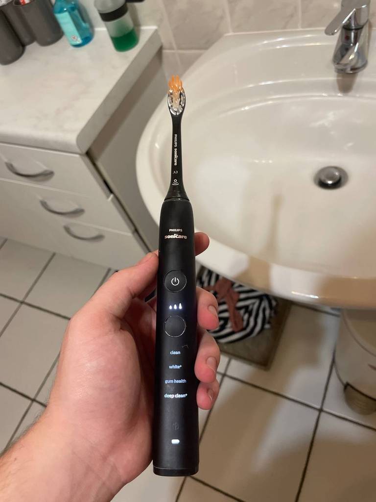 Eine schwarze, getestete Schallzahnbürste des Typs Philips Sonicare befindet sich in einer linken Hand im Waschbeckenbereich.