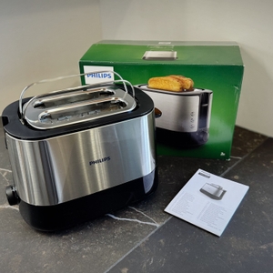 Der getestete Toaster von Philips steht mit der Verpackung und der Bedienungsanleitung auf einer dunklen Arbeitsfläche.