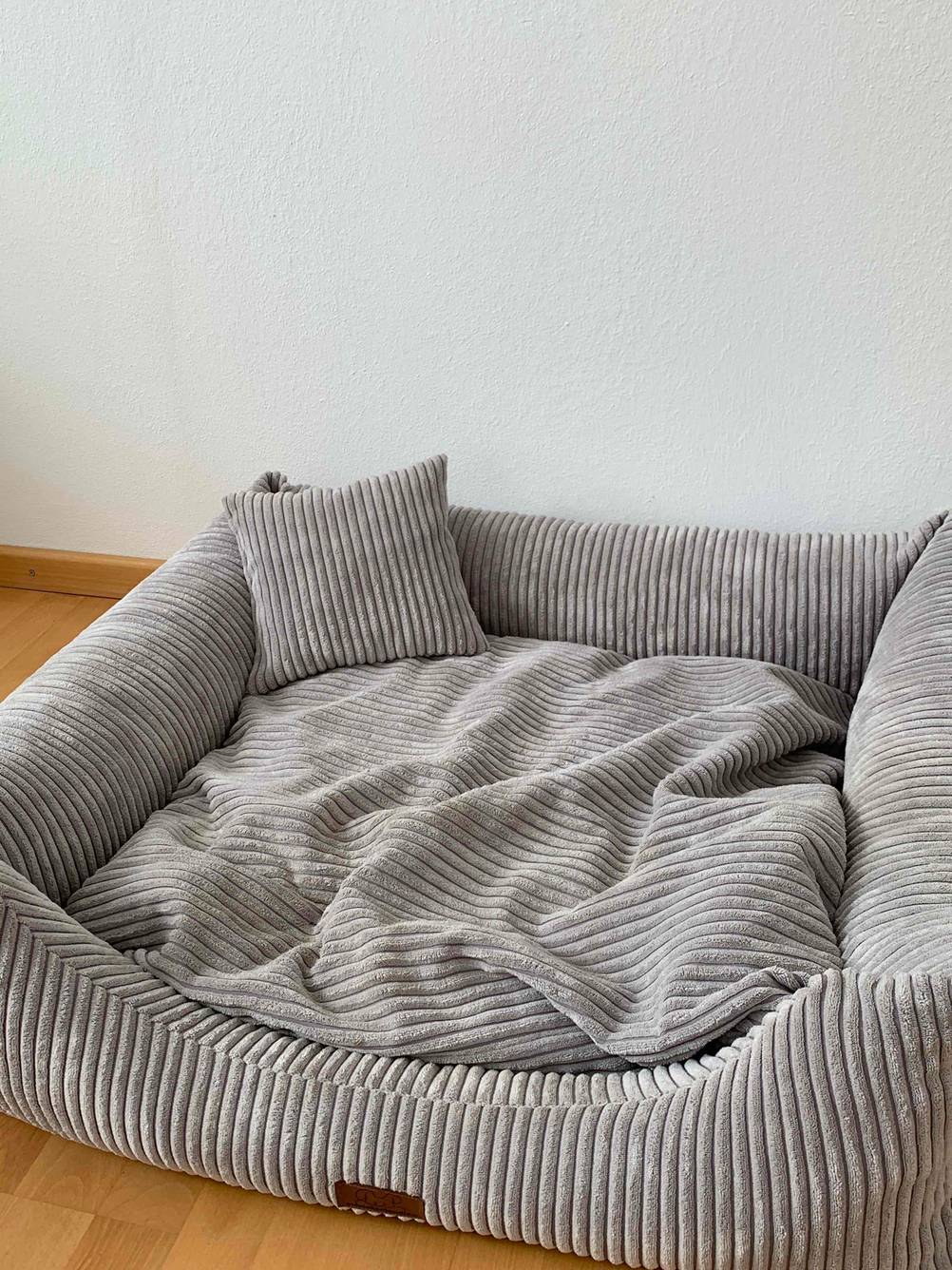 Ein graues Hundebett mit Kissen steht auf dem Boden.
