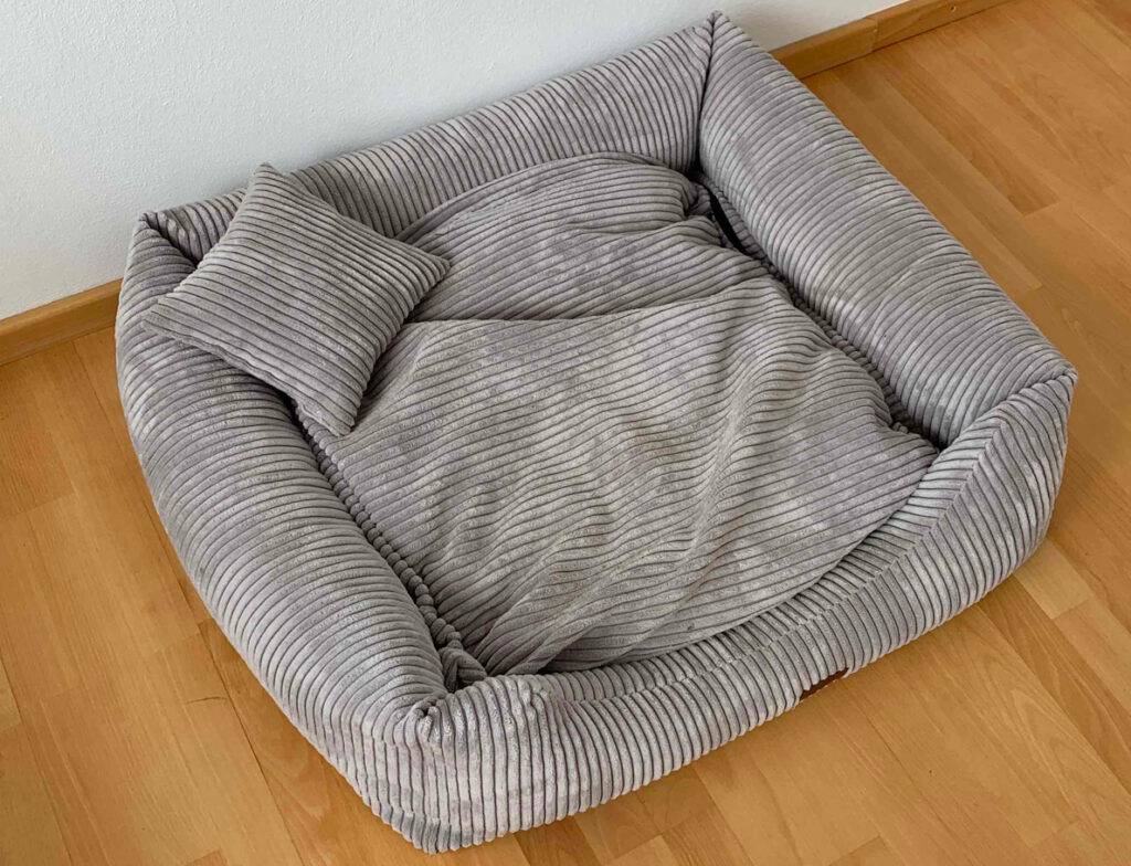 Ansicht von oben auf eine hellgraues Hundebett im Test, das auf einem Parkettboden an der weißen Wand platziert ist.